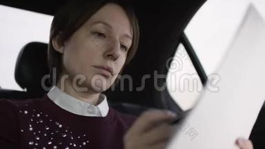 穿着白色上衣和栗色毛衣的棕色头发的漂亮女人坐在车上，学习工作文件